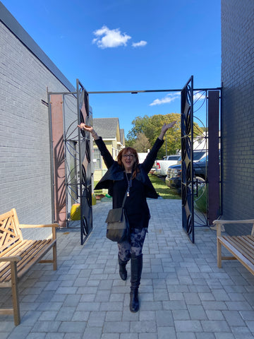 Robin Baron raises her arms as she walks through a large, decorative metal doorway with a blue sky in the background.