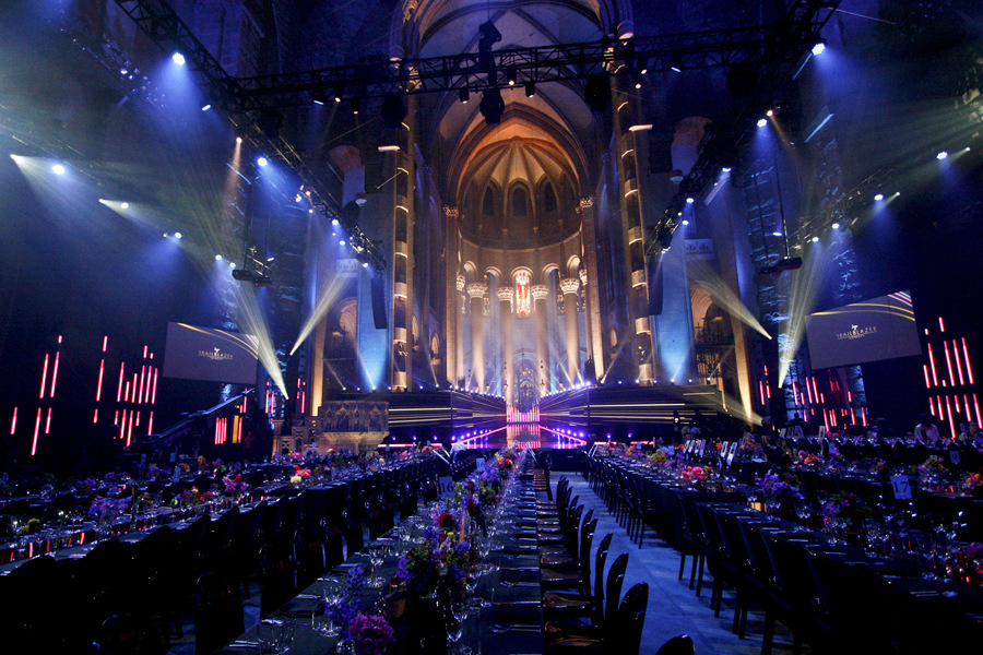 This pictures what looks like a grand awards show or major event. Purple colored lights dimly light long rows of elegantly set tables, with flowers running down their center. The backdrop a stage with that looks like a grand cathedral, with a curved dome roof, columns, and stained glass windows. Moving lighting fixtures at either side send cascading spotlights in angled directions from the sleek black stage itself