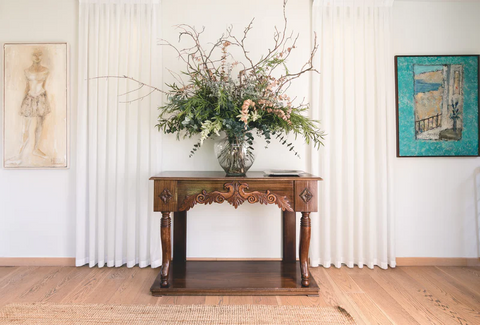 Decorative wooden table with a vase and many flowers on it. Framed on either side by curtains and pieces of art