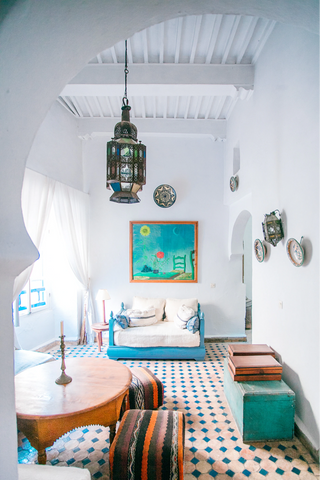 A room decorated in bright colors, with a blue diamond patterned rug, a bright blue couch frame with white cushions, a teal-dominant painting on the back wall. Light pours in from a window on the left side. Other furniture includes a side table with a lamp, a table with a candlestick, a bench on the right wall with decorative wall decor hanging above it.